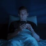 Mann scrollt nachts im Bett im bläulichen Licht seines Smartphones
