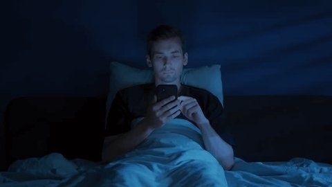 Mann scrollt nachts im Bett im bläulichen Licht seines Smartphones